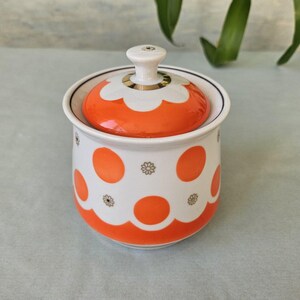 Könnte beinhalten: Eine weiße Zuckerdose aus Keramik mit orangefarbenen Punkten und einem passenden Deckel. Der Deckel hat einen weißen Griff und ein Blumenmuster. Ein Vintage-Küchenartikel.