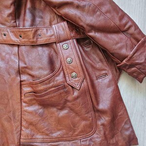 May include: A brown leather jacket with a belt and pocket detail. The jacket has a classic design with a buttoned closure and a tailored fit. The leather appears to be well-worn, giving it a vintage look. The jacket is a medium brown color.