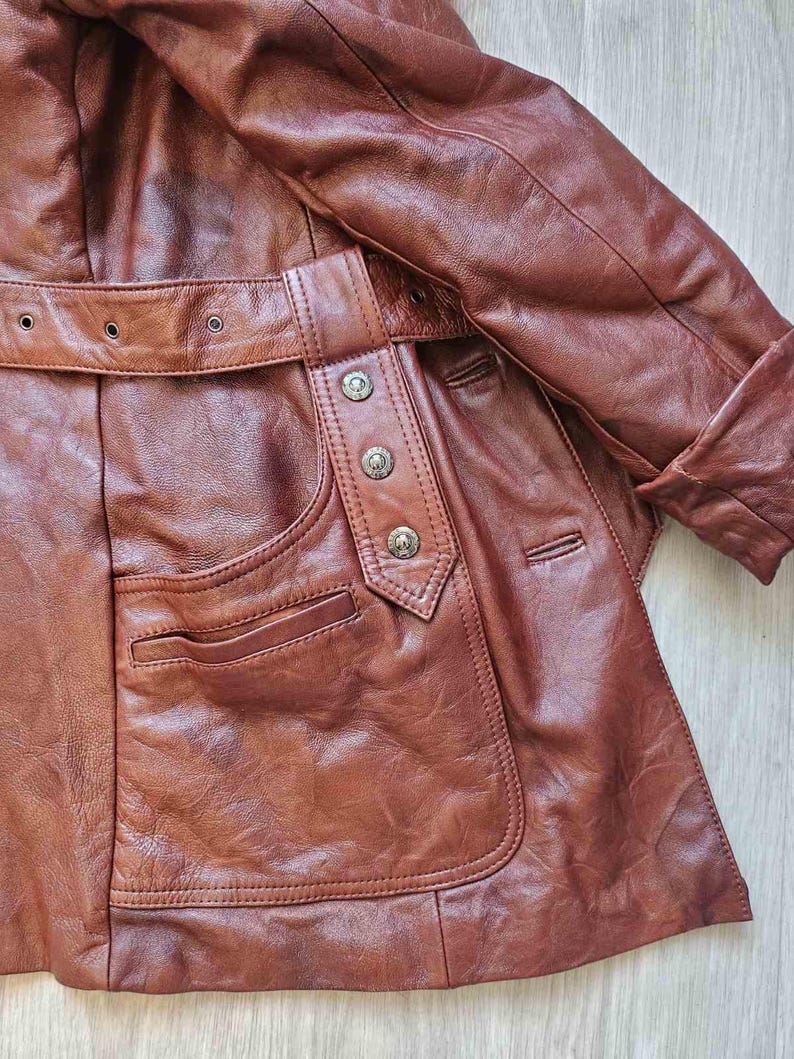 May include: A brown leather jacket with a belt and pocket detail. The jacket has a classic design with a buttoned closure and a tailored fit. The leather appears to be well-worn, giving it a vintage look. The jacket is a medium brown color.