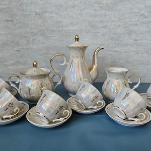 May include: A pearlescent tea set with gold trim, including a teapot, creamer, sugar bowl, and six teacups with saucers. The set has a delicate floral design and is arranged on a blue surface.