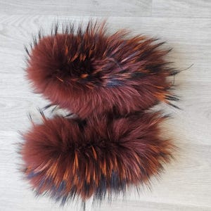 May include: Two fluffy, reddish-brown fur pieces with black and orange highlights. The fur appears soft and dense, likely from an animal. The pieces are arranged side-by-side on a light gray wooden surface.