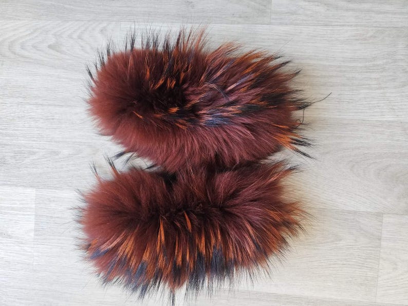 May include: Two fluffy, reddish-brown fur pieces with black and orange highlights. The fur appears soft and dense, likely from an animal. The pieces are arranged side-by-side on a light gray wooden surface.