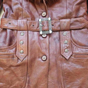 May include: A brown leather jacket with a fur collar and a belt. The jacket has a decorative silver buckle and silver button details. The pockets are outlined with stitching. The jacket is a medium brown color.