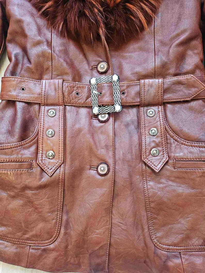 May include: A brown leather jacket with a fur collar and a belt. The jacket has a decorative silver buckle and silver button details. The pockets are outlined with stitching. The jacket is a medium brown color.