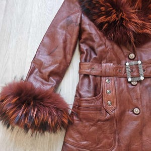 May include: A brown leather jacket with a fur collar and cuffs. The jacket features a belt with a decorative buckle and button details. The fur trim has shades of red, orange, and black.