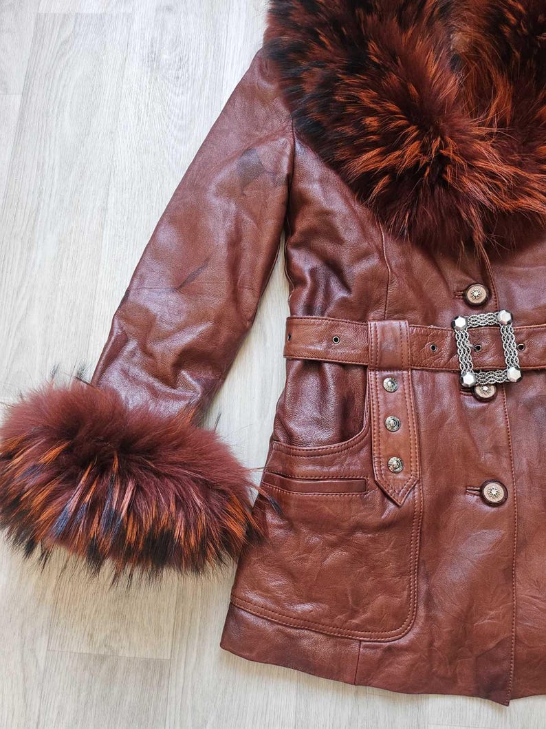 May include: A brown leather jacket with a fur collar and cuffs. The jacket features a belt with a decorative buckle and button details. The fur trim has shades of red, orange, and black.