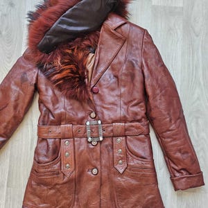 May include: A brown leather coat with a fur-trimmed hood and belt. The coat features a button closure, pockets, and a decorative buckle on the belt. The fur trim is a mix of red, orange, and black.