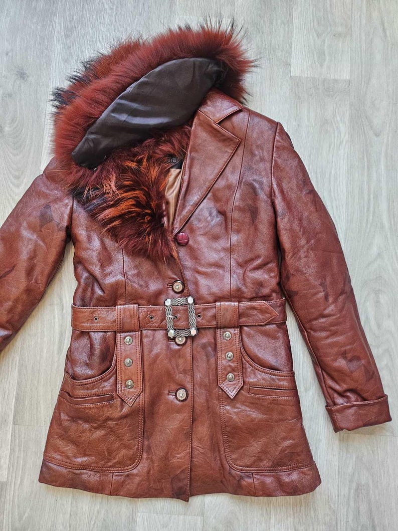May include: A brown leather coat with a fur-trimmed hood and belt. The coat features a button closure, pockets, and a decorative buckle on the belt. The fur trim is a mix of red, orange, and black.