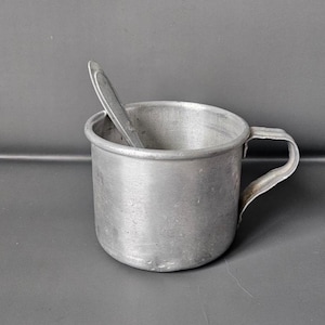 May include: A small, silver-colored metal cup with a handle, holding a spoon. The cup has a rounded shape and a simple design. The spoon is also silver and rests inside the cup. The cup and spoon are set against a gray background.