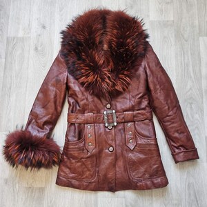 May include: A brown leather coat with a fur collar and cuffs. The coat features a belted waist with a decorative buckle, button closures, and pockets. The fur trim is a mix of brown and reddish-brown tones.