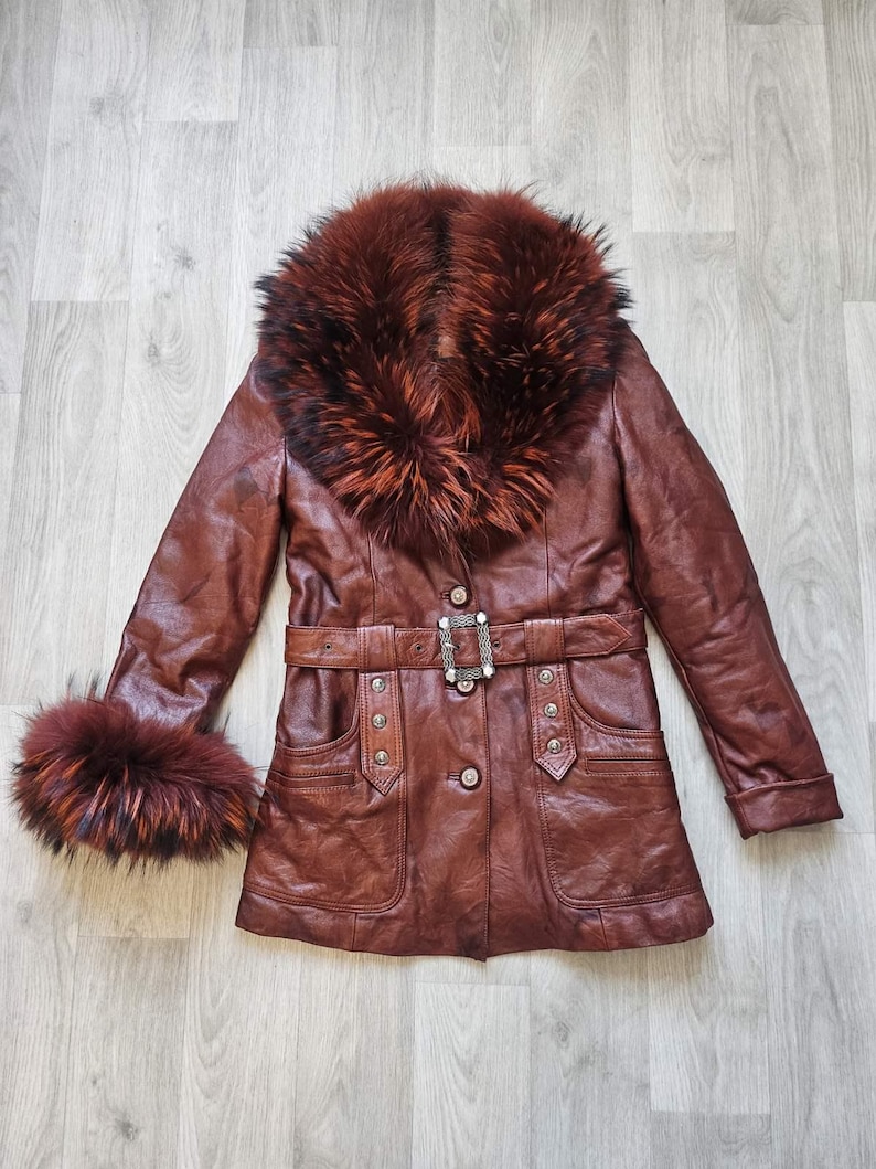 May include: A brown leather coat with a fur collar and cuffs. The coat features a belted waist with a decorative buckle, button closures, and pockets. The fur trim is a mix of brown and reddish-brown tones.