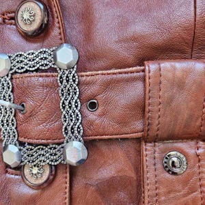 May include: Close-up of a brown leather jacket with decorative silver-toned metal buttons and a buckle. The jacket features detailed stitching and a textured surface, showcasing a vintage aesthetic.