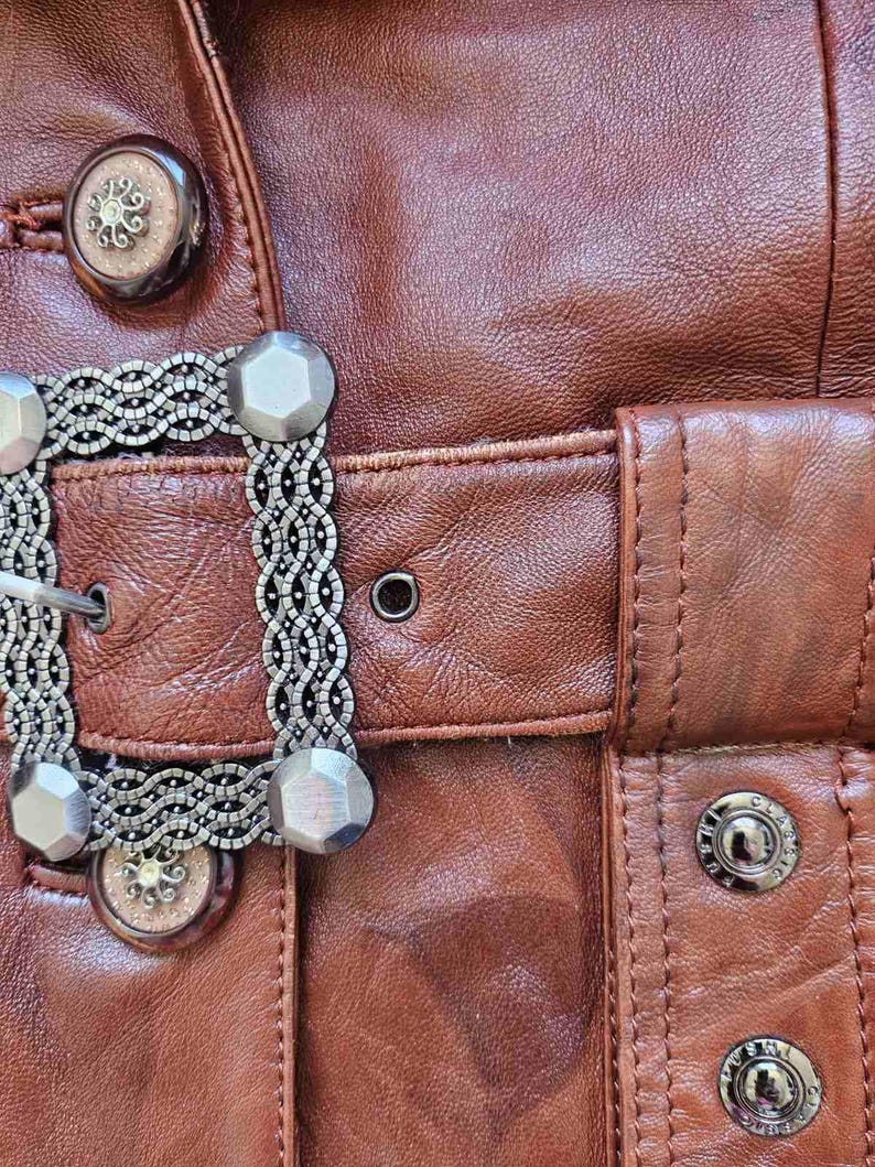 May include: Close-up of a brown leather jacket with decorative silver-toned metal buttons and a buckle. The jacket features detailed stitching and a textured surface, showcasing a vintage aesthetic.