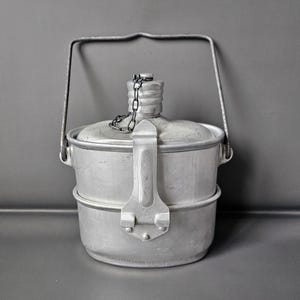 May include: A vintage, silver-colored canteen with a handle and a hinged clasp. The canteen has a cylindrical shape with a screw-on cap attached by a chain. The canteen is made of metal and appears to be well-used.