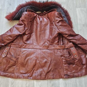 May include: A brown leather coat with a fur-trimmed collar. The coat features a belted waist, two front pockets, and decorative buttons. The leather has a rich, textured appearance, and the coat is laid out on a light-colored wooden surface.