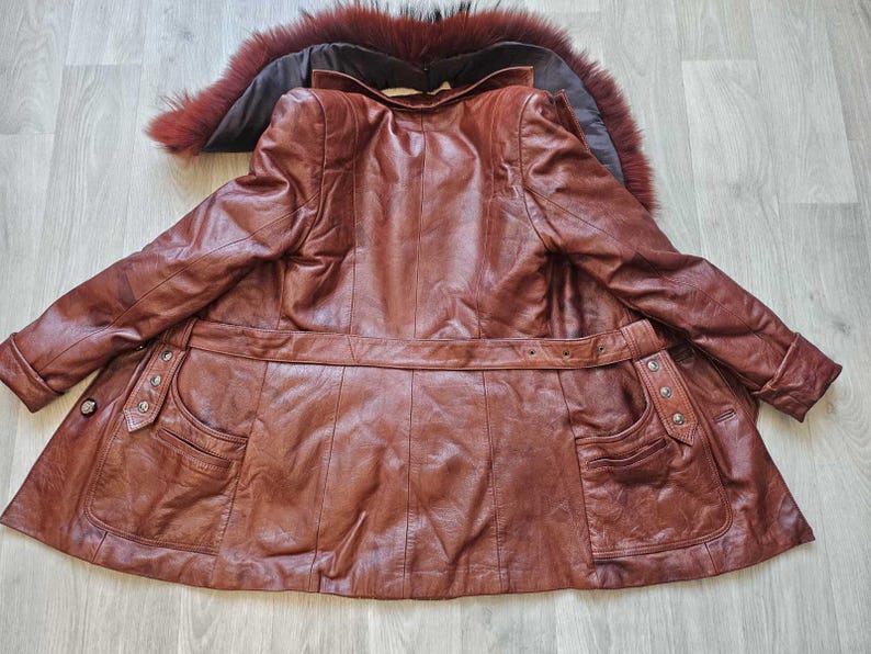 May include: A brown leather coat with a fur-trimmed collar. The coat features a belted waist, two front pockets, and decorative buttons. The leather has a rich, textured appearance, and the coat is laid out on a light-colored wooden surface.