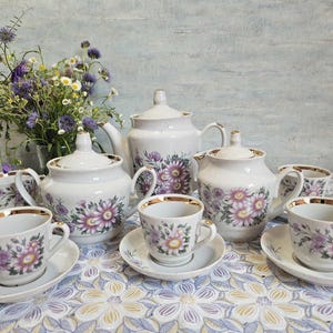 May include: A white porcelain tea set with a floral design featuring purple and yellow flowers. The set includes a teapot, sugar bowl, creamer, and several teacups and saucers, all with gold trim.