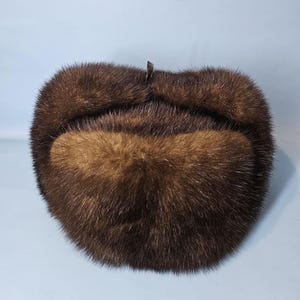 Puede incluir: Un sombrero de piel marrón con forma redondeada y centro marrón oscuro. El sombrero tiene un aspecto suave y texturizado y un pequeño cierre metálico en la parte superior. El sombrero está diseñado para cubrir las orejas y la parte posterior del cuello.