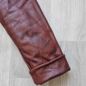 May include: Close-up of a brown leather jacket sleeve with a folded cuff. The leather has a textured appearance and a reddish-brown hue. The cuff is neatly folded, showcasing the jacket's tailoring.