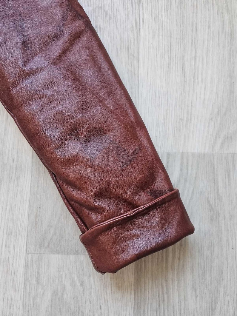 May include: Close-up of a brown leather jacket sleeve with a folded cuff. The leather has a textured appearance and a reddish-brown hue. The cuff is neatly folded, showcasing the jacket's tailoring.