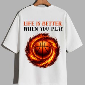 May include: White t-shirt featuring a basketball engulfed in flames. The shirt has the text "LIFE IS BETTER WHEN YOU PLAY" in orange and black. The design is centered on the front of the shirt.