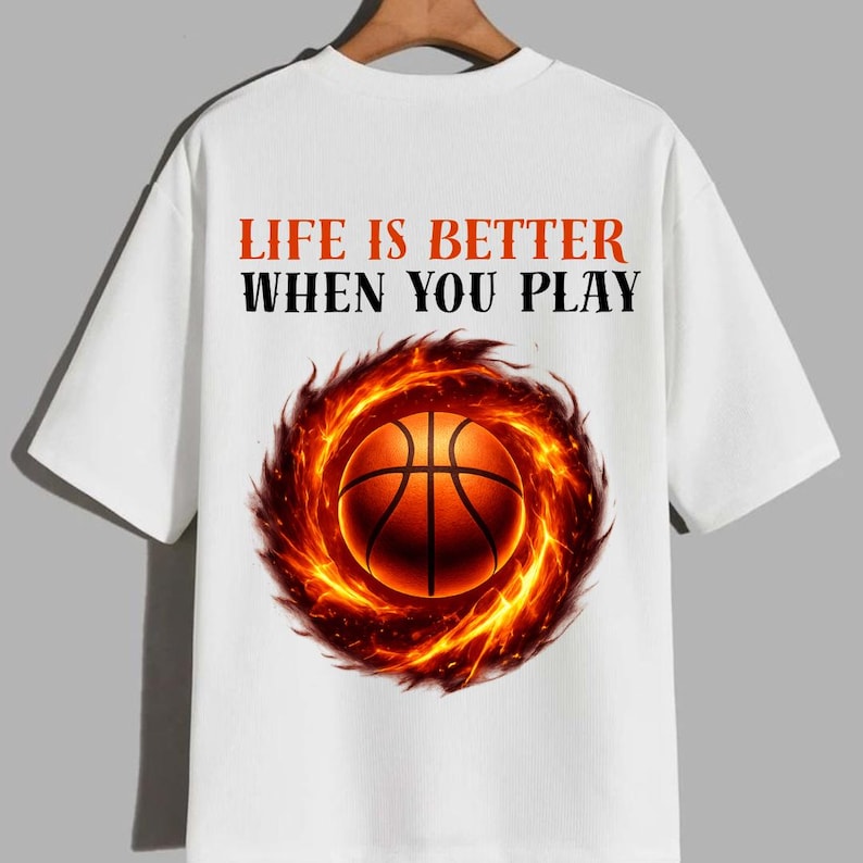 May include: White t-shirt featuring a basketball engulfed in flames. The shirt has the text "LIFE IS BETTER WHEN YOU PLAY" in orange and black. The design is centered on the front of the shirt.