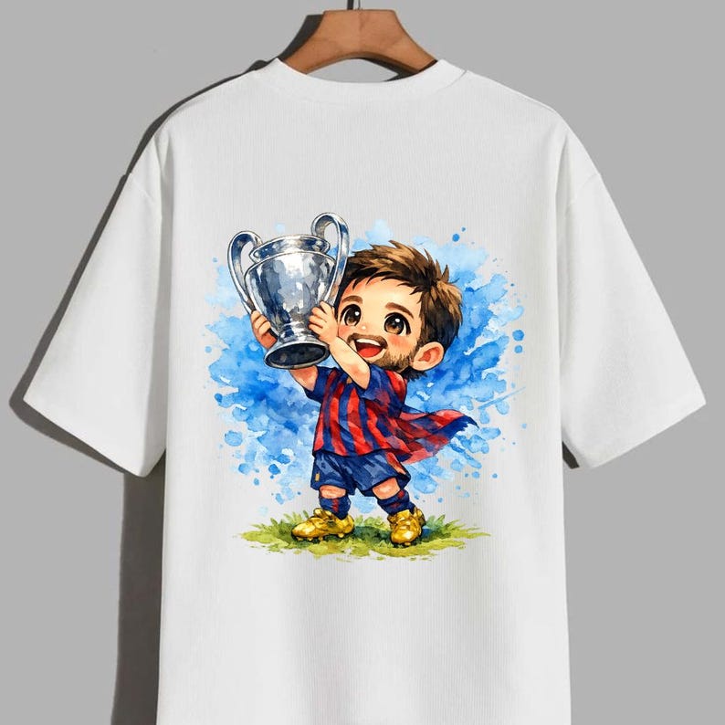 May include: White t-shirt featuring a cartoon illustration of a soccer player holding a trophy. The player is depicted with a red and blue striped uniform, blue shorts, and gold shoes, set against a blue watercolor background.