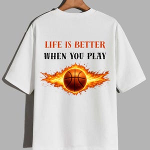 May include: White t-shirt featuring a basketball design. The basketball is surrounded by flames, with the text "LIFE IS BETTER WHEN YOU PLAY" in red and black. The shirt has short sleeves and a crew neck.