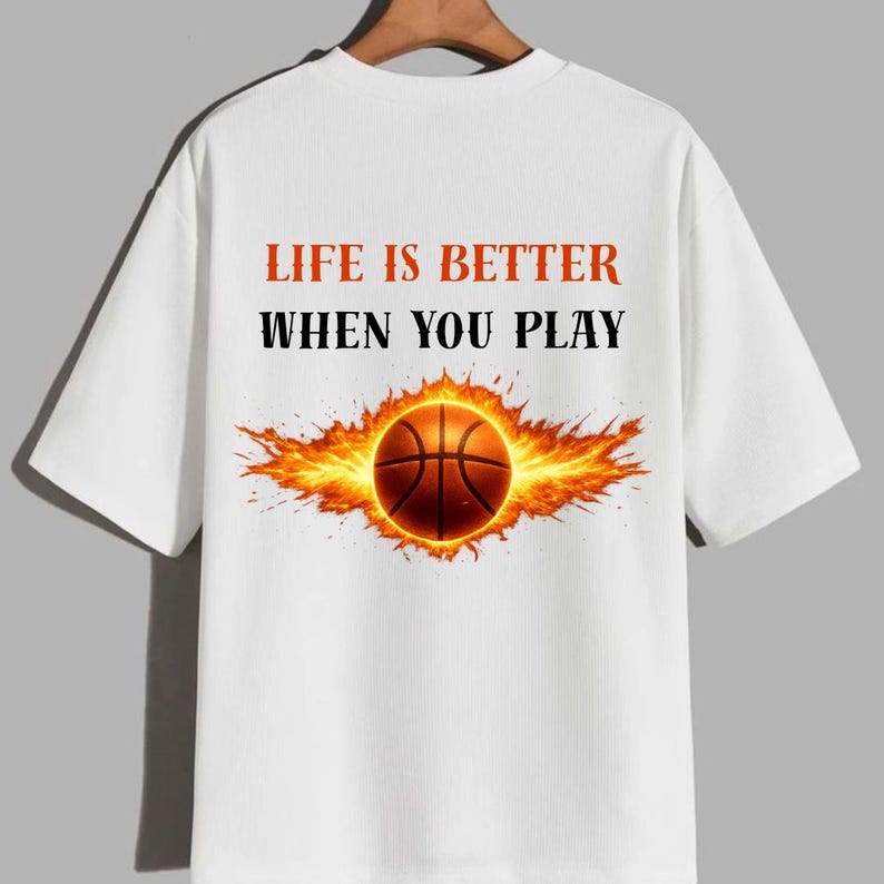 May include: White t-shirt featuring a basketball design. The basketball is surrounded by flames, with the text "LIFE IS BETTER WHEN YOU PLAY" in red and black. The shirt has short sleeves and a crew neck.