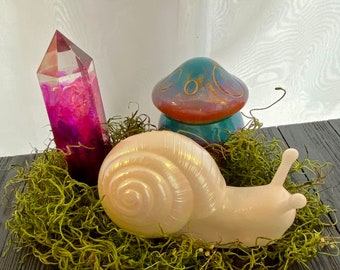 Puede incluir: Una figurita de caracol de cerámica blanca con un acabado nacarado se sienta sobre un lecho de musgo verde. También se puede ver un cristal rosa y un hongo de cerámica azul y marrón en la imagen.