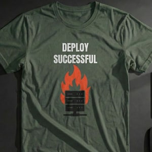 May include: Olive green t-shirt with the words "DEPLOY SUCCESSFUL" in white. A graphic of a server on fire is below the text. The shirt is displayed on a dark surface with a keyboard, laptop, mouse, and other items.
