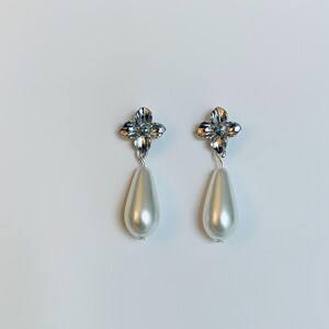 Peut inclure: Une paire de boucles d'oreilles pendantes. Chaque boucle d'oreille pr&eacute;sente un motif de fleur &agrave; quatre p&eacute;tales de couleur argent&eacute;e en haut, reli&eacute; &agrave; une fausse perle en forme de poire, blanc cass&eacute;. Les boucles d'oreilles sont pos&eacute;es sur un fond blanc.
