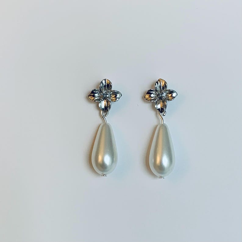 Peut inclure: Une paire de boucles d'oreilles pendantes. Chaque boucle d'oreille pr&eacute;sente un motif de fleur &agrave; quatre p&eacute;tales de couleur argent&eacute;e en haut, reli&eacute; &agrave; une fausse perle en forme de poire, blanc cass&eacute;. Les boucles d'oreilles sont pos&eacute;es sur un fond blanc.