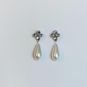 Peut inclure: Une paire de boucles d'oreilles &eacute;l&eacute;gantes avec un motif floral argent&eacute; et une perle de fausse imitation en forme de goutte, de couleur blanc cass&eacute;. Le fond est blanc.