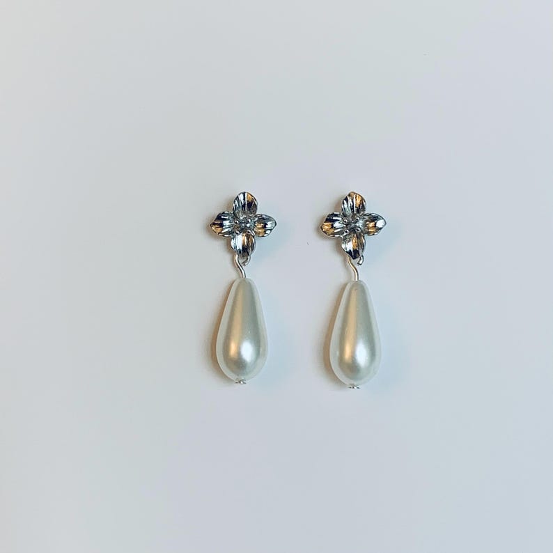 Peut inclure: Une paire de boucles d'oreilles &eacute;l&eacute;gantes avec un motif floral argent&eacute; et une perle de fausse imitation en forme de goutte, de couleur blanc cass&eacute;. Le fond est blanc.
