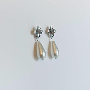 Peut inclure: Une paire de boucles d'oreilles pendantes &eacute;l&eacute;gantes. Chaque boucle d'oreille est orn&eacute;e d'un clou floral argent&eacute; et d'une fausse perle blanche en forme de goutte. Les boucles d'oreilles sont con&ccedil;ues pour pendre, cr&eacute;ant un look classique et sophistiqu&eacute;.