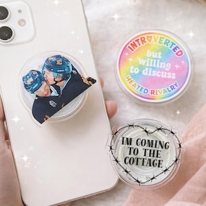 Puede incluir: Una funda transparente para teléfono con un soporte para teléfono con temática de hockey que presenta a dos jugadores con uniformes azules y negros. Se ven dos soportes para teléfono adicionales, uno con texto arcoíris que dice "Introvertido pero dispuesto a discutir la rivalidad acalorada."