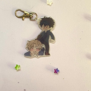 Op de afbeelding: Een heldere acryl sleutelhanger met twee anime-stijl karakters. Eén karakter staat en de andere ligt op de grond. De sleutelhanger heeft een goudkleurige sluiting. Verspreid rond de sleutelhanger zijn kleine stervormige decoraties in verschillende kleuren.