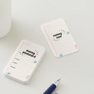 May include: Two white notepads with lined pages and the phrases "missing photocard" and "coming soon" in black text. Each notepad has pink dots along the left edge and blue star and exclamation point illustrations. A blue pen and a white object are also visible.