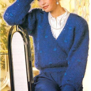 May include: A woman wearing a blue sweater with a V-neck and blue polka dots. She is wearing a white blouse and a pearl necklace. She is sitting on a black chair.