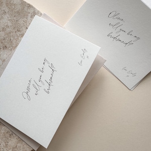 May include: Two white cards with elegant calligraphy asking "Jessica, will you be my bridesmaid?" and "Olivia, will you be my bridesmaid?" The cards are signed "Love Emily". The cards are on a neutral background.
