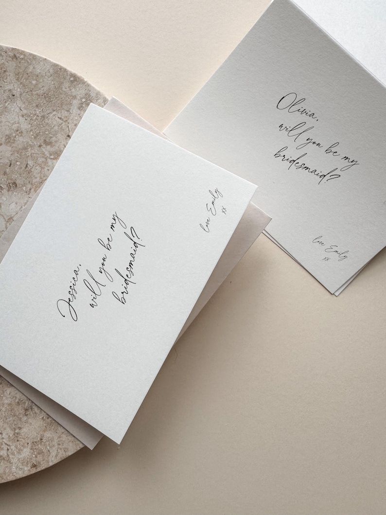May include: Two white cards with elegant calligraphy asking "Jessica, will you be my bridesmaid?" and "Olivia, will you be my bridesmaid?" The cards are signed "Love Emily". The cards are on a neutral background.