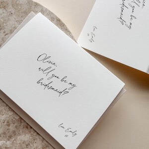 May include: Two white cards with the text "Olivia, will you be my bridesmaid?" and "Jessica, will you be my bridesmaid?" in elegant script. Each card is signed "Love Emily" with two kisses. The cards are on a neutral background.