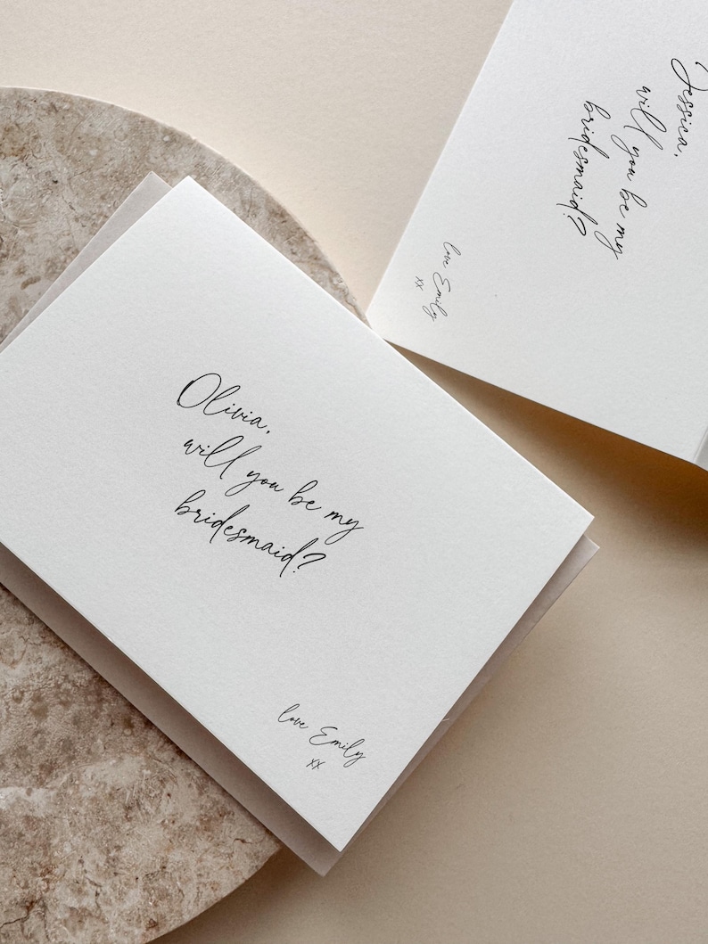 May include: Two white cards with the text "Olivia, will you be my bridesmaid?" and "Jessica, will you be my bridesmaid?" in elegant script. Each card is signed "Love Emily" with two kisses. The cards are on a neutral background.