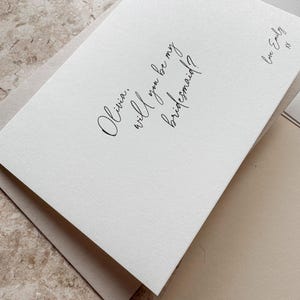 May include: A white card with the text "Olivia, will you be my bridesmaid?" in elegant script. The card is layered over a neutral-toned envelope, with a signature that reads "Love Emily".