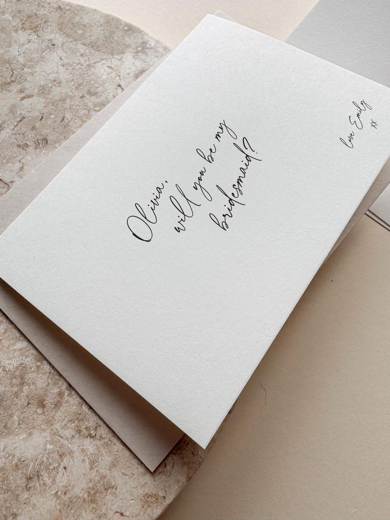 May include: A white card with the text "Olivia, will you be my bridesmaid?" in elegant script. The card is layered over a neutral-toned envelope, with a signature that reads "Love Emily".