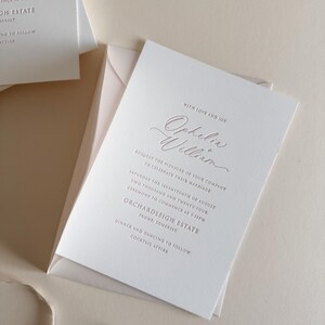 May include: Elegant wedding invitation with the names "Ophelia William" in a delicate script. The invitation is white with rose gold text and is placed inside a matching envelope. The text includes the date, time, and location of the ceremony.