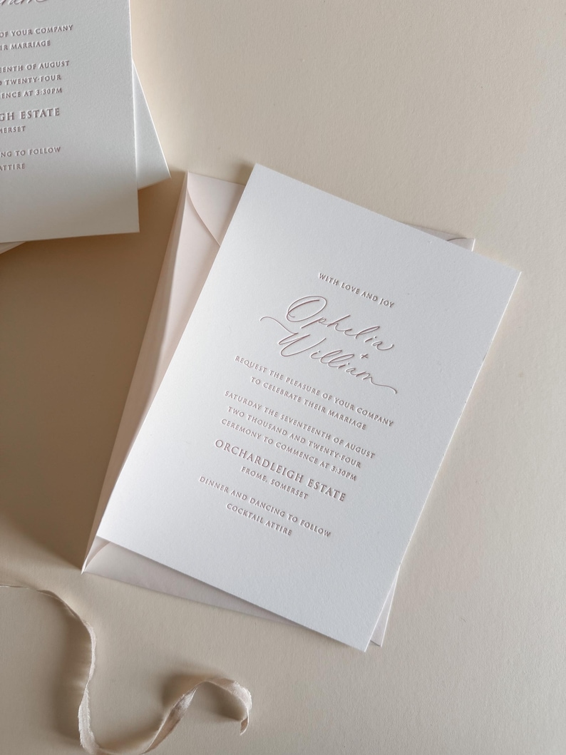 May include: Elegant wedding invitation with the names "Ophelia William" in a delicate script. The invitation is white with rose gold text and is placed inside a matching envelope. The text includes the date, time, and location of the ceremony.