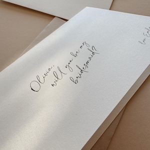 May include: Close-up of ivory-colored cards with handwritten calligraphy. The cards read "Olivia, will you be my bridesmaid?" and "Jessica, will you be my bridesmaid?" with a signature. The cards are stacked on a neutral-toned surface.
