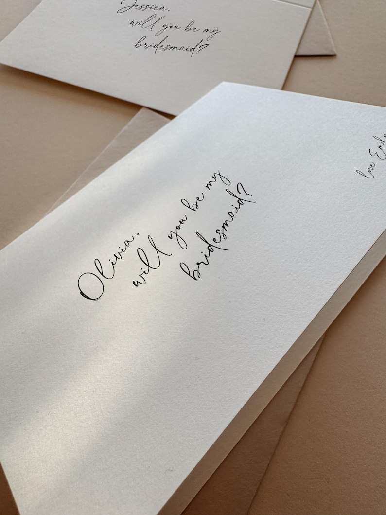 May include: Close-up of ivory-colored cards with handwritten calligraphy. The cards read "Olivia, will you be my bridesmaid?" and "Jessica, will you be my bridesmaid?" with a signature. The cards are stacked on a neutral-toned surface.